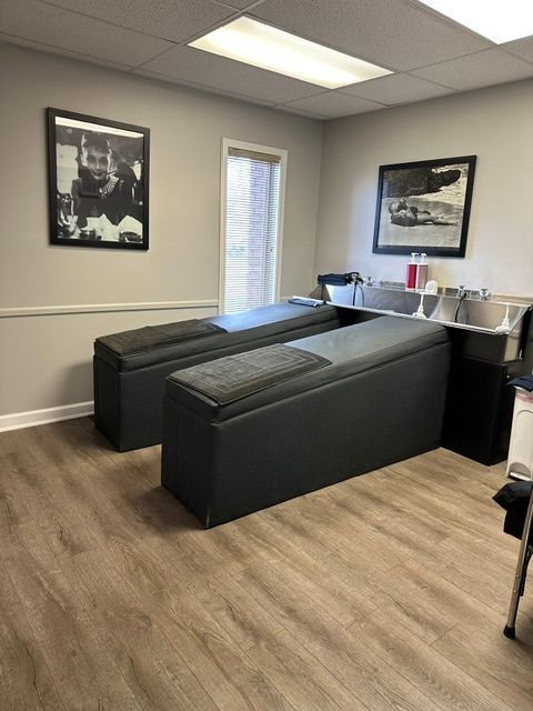Two gray massage tables placed side-by-side in a room with light wood floors, a window, and framed art on the walls.