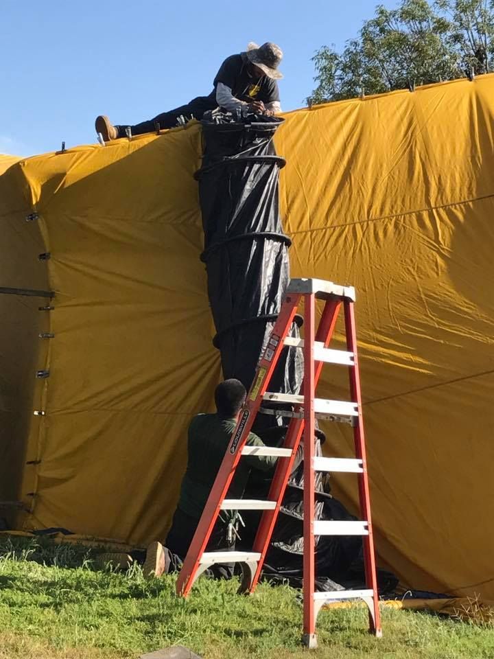 Two workers install a black chute onto a large, yellow fabric structure outdoors.