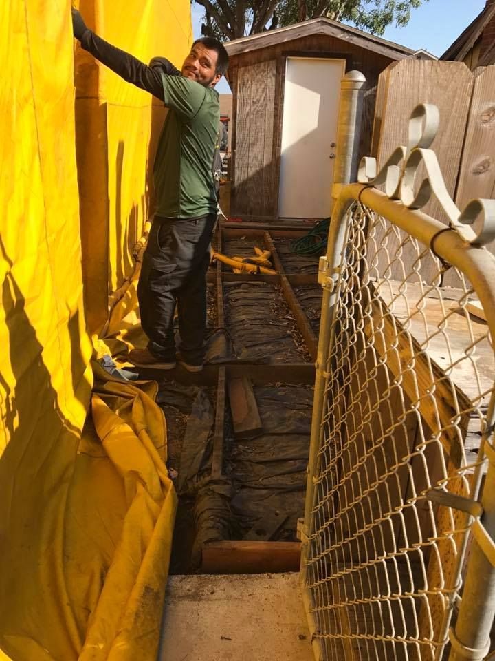 A person in a green shirt reaches toward a yellow wall near a chain-link fence and a small shed in an outdoor work area.