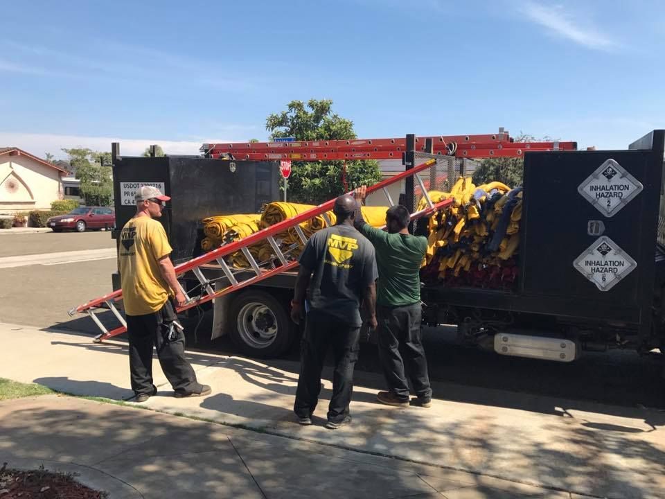 Three people work to load yellow industrial equipment and a long red ladder onto the back of a black utility truck.
