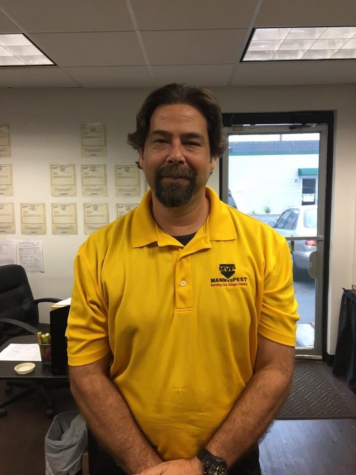 A person with a beard wearing a yellow polo shirt with a logo, standing in an office with certificates on the wall.