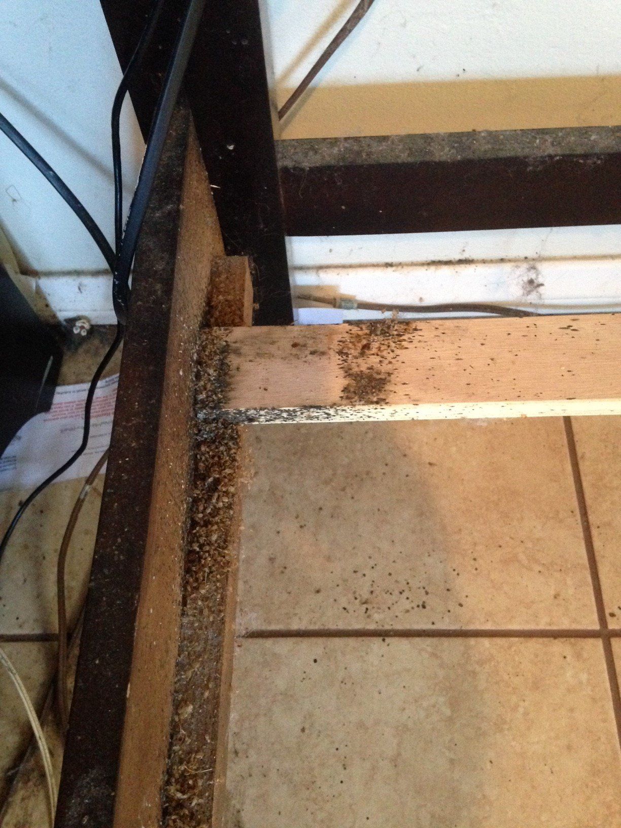 Close-up of accumulated dark debris and wood dust at the corner joint of a piece of particleboard furniture on a tile floor.