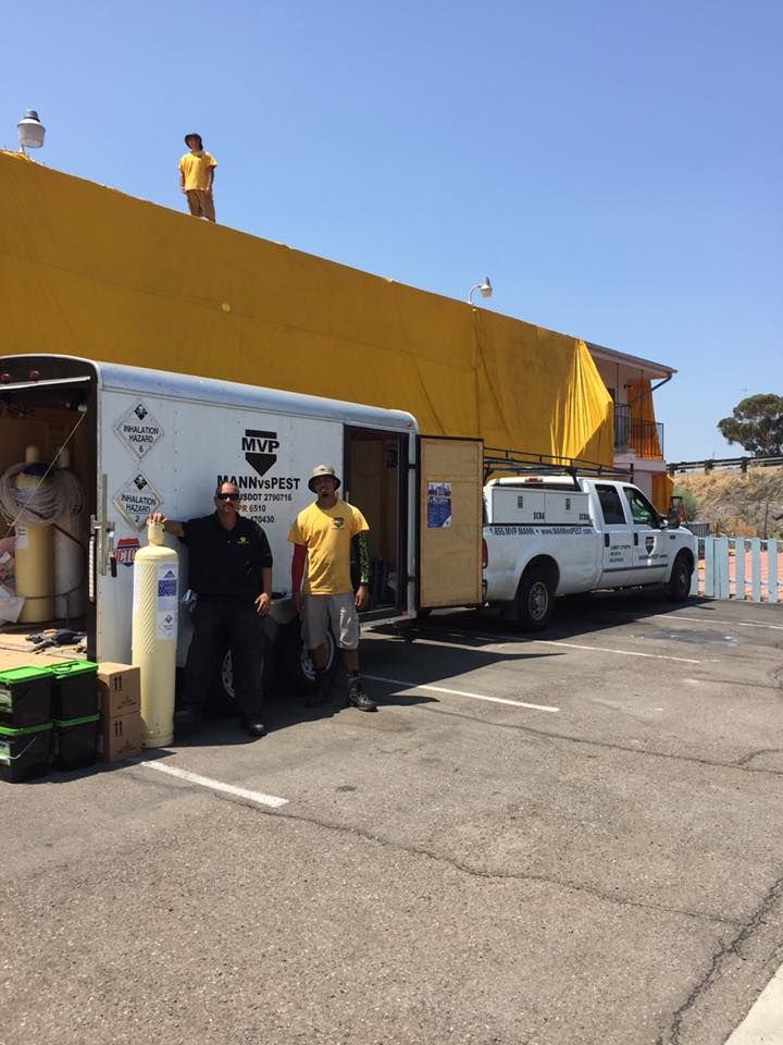 Two workers stand by a white trailer while another is on the roof of a building covered in yellow tarps.
