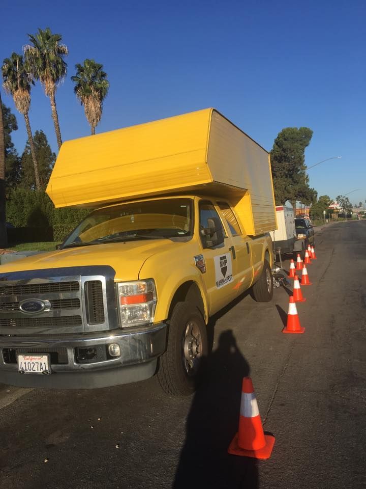 A bright yellow utility truck with a large, matching cargo box is parked on a road beside a line of orange traffic cones.