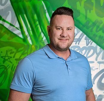 Man with dark hair and a blue polo shirt smiles in front of a green graffiti wall.