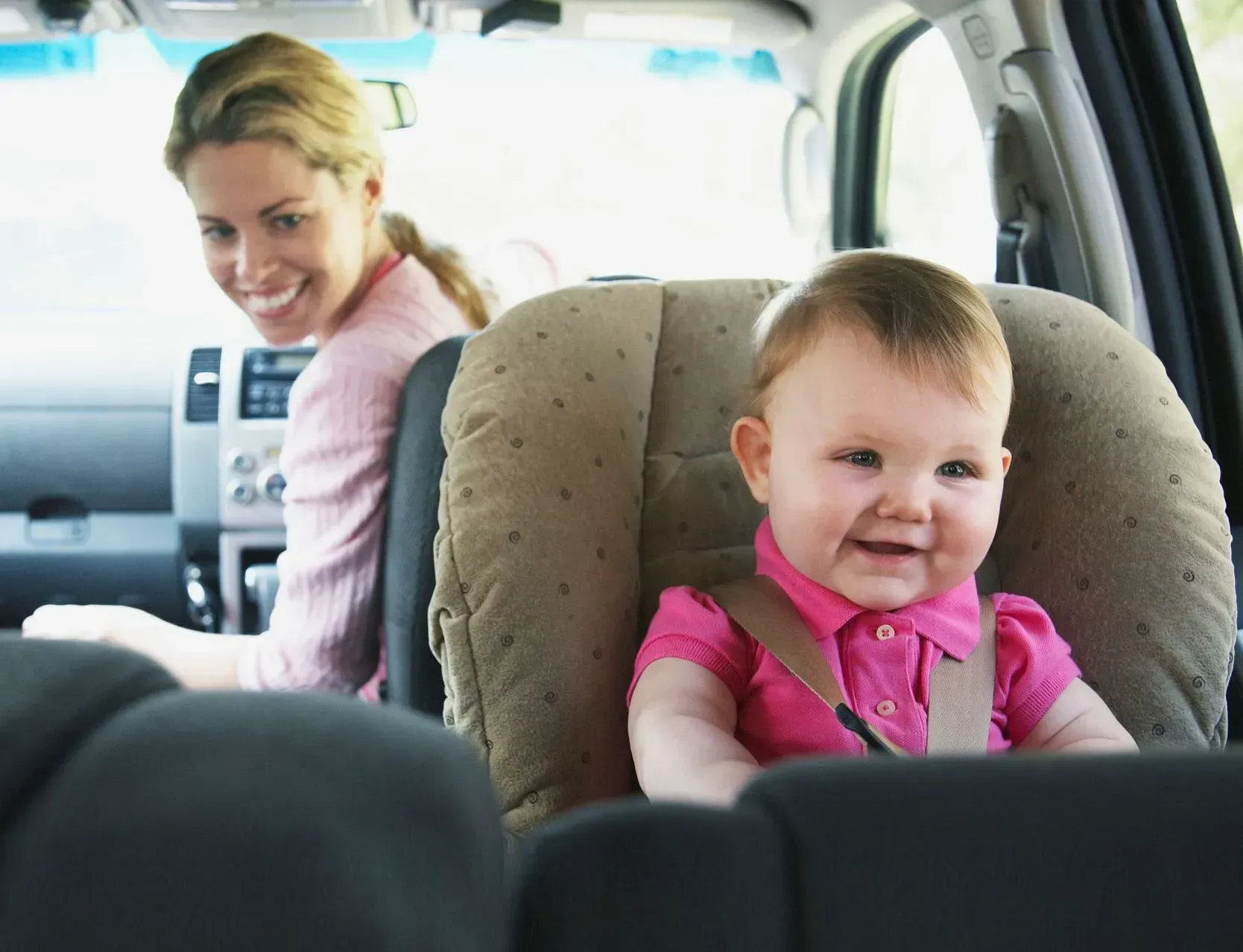 Smiling baby in car seat, woman in driver's seat. Pink clothing, vehicle interior.