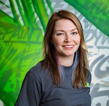 Woman with long brown hair smiling, wearing a gray t-shirt, standing in front of graffiti.