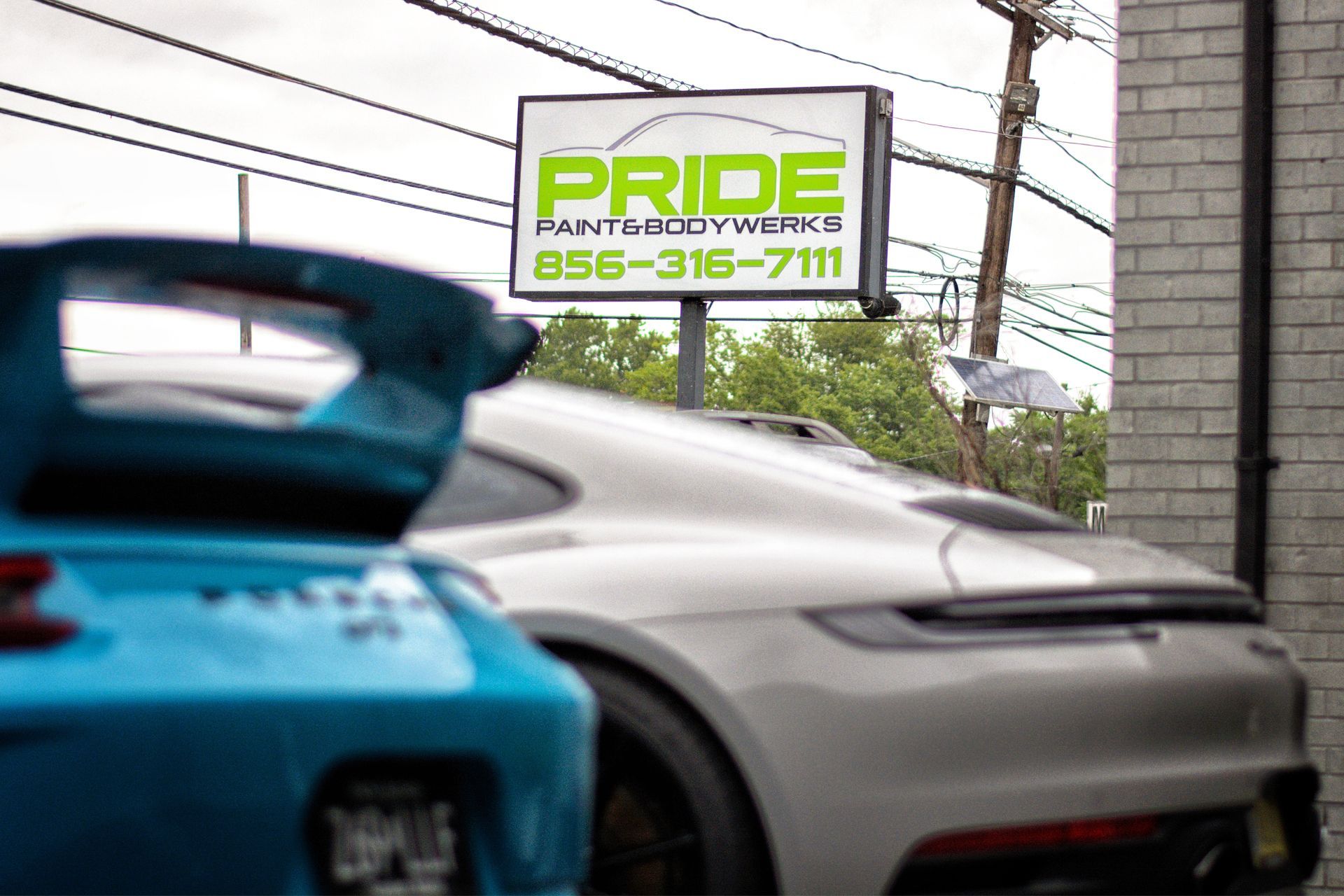 Two Porsche sports cars parked in front of Pride Paint & Body Works sign; light blue and silver.