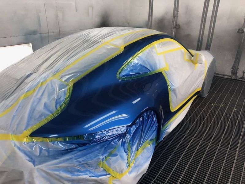 Blue sports car partially covered with plastic and taped in a paint booth.