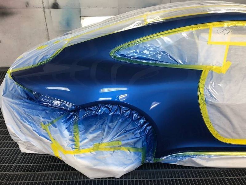 Blue car body panel partially masked with plastic and tape for painting in a spray booth.