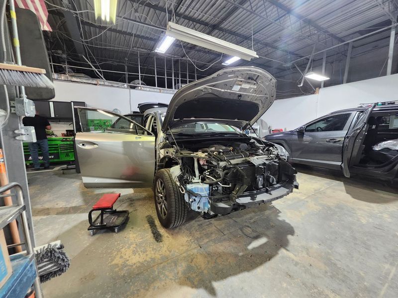 Car in a repair shop with hood and door open, front end damaged. Tools and a person visible.