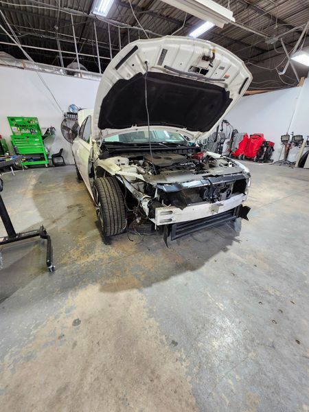 White car with open hood in a repair shop, front damaged, awaiting repair.