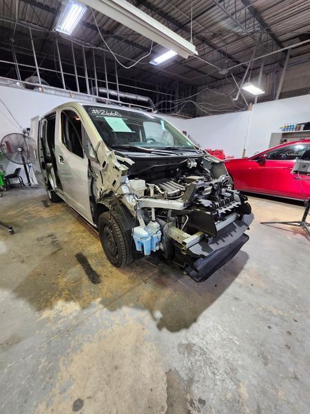 Damaged silver van in a repair shop with its front end disassembled, next to a red car.