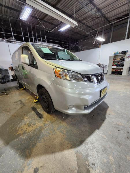Silver Nissan NV200 van inside a garage. The van is on jacks, parked near a white wall.