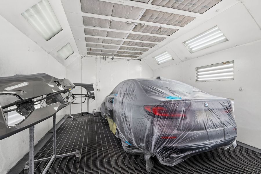 Car in a paint booth covered in plastic; car parts on a stand to the left.
