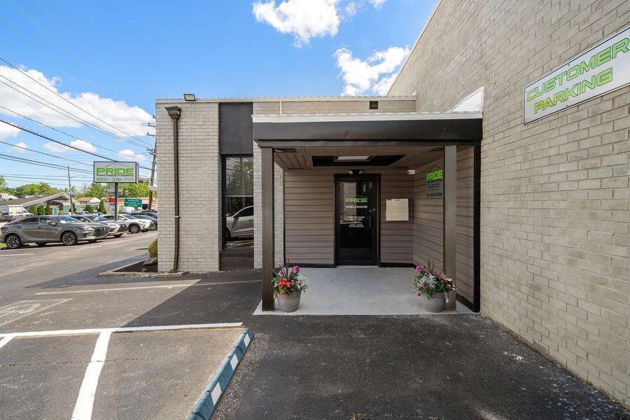 Entrance to a business with light-colored siding, a black door, and flowers. Parking spaces and street view.