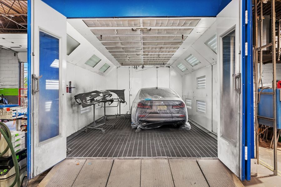 Car inside a paint booth with open doors; white walls, blue trim, and gray floor.