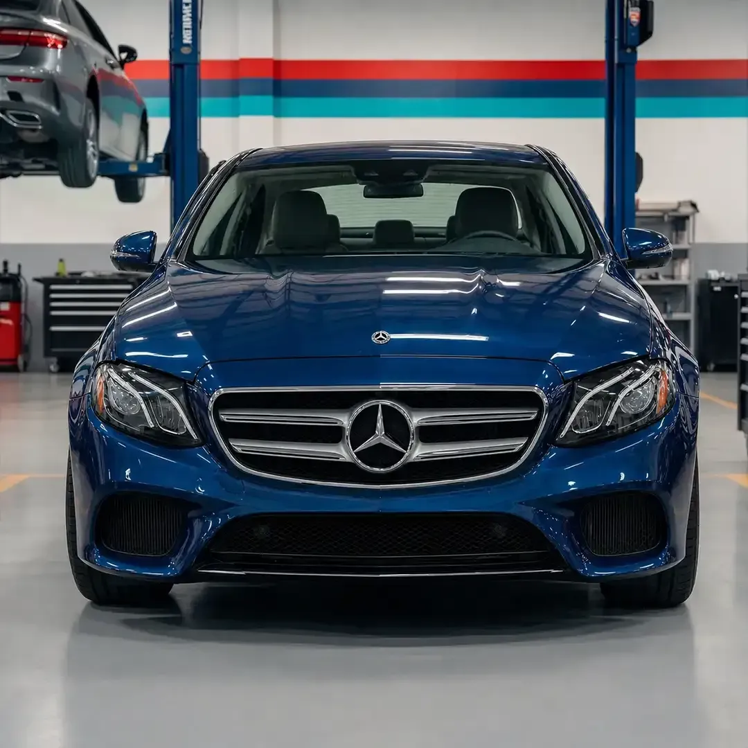 Front view of a blue Mercedes sedan in an auto workshop, with a lifted car in the background.
