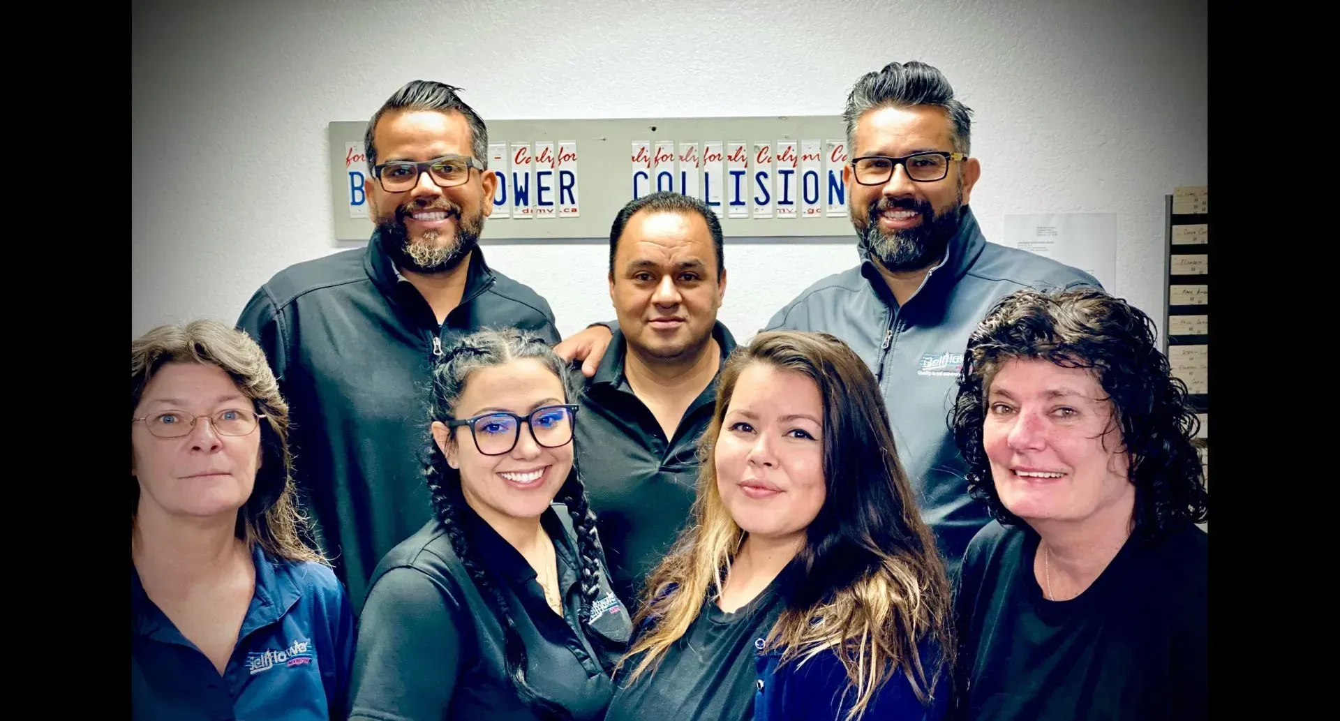 Seven people smiling in a group photo indoors, standing in front of a sign.