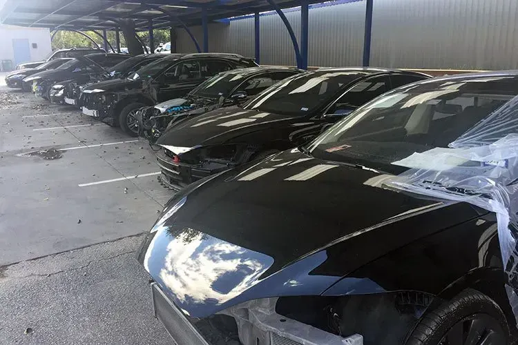 Several black cars parked in a covered lot, front car damaged with a crumpled hood and headlight area