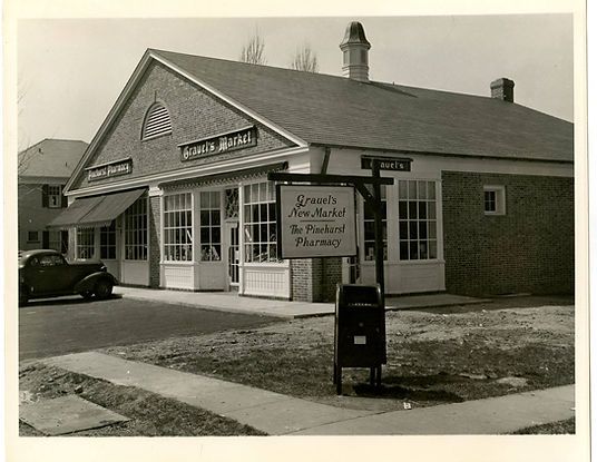 Black-and-white photo of a small brick storefront with a sign and a parked car beside the entrance