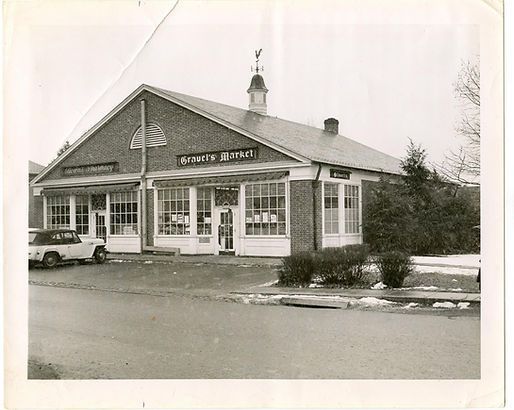 Black-and-white photo of a small corner shop with a parked car and storefront windows