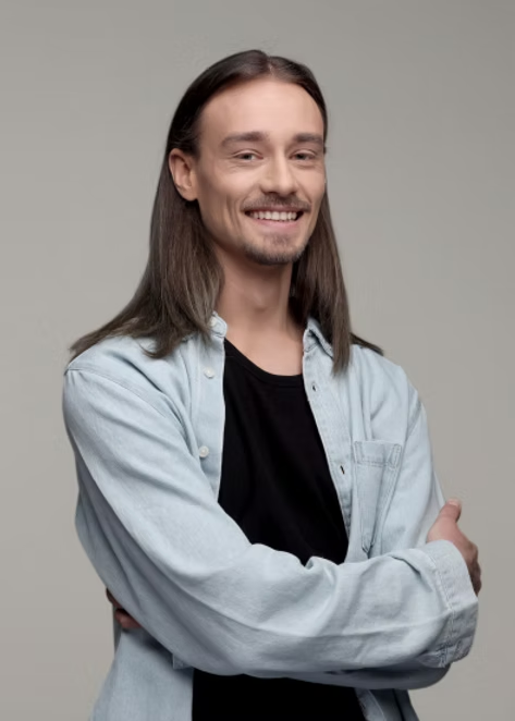 Smiling person with long hair in a light blue shirt, arms crossed against a plain gray background