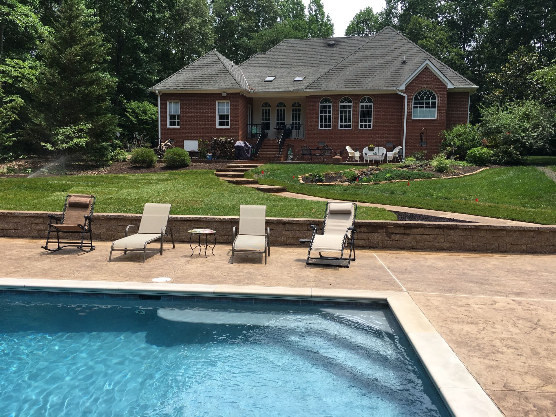 Backyard with pool, lounge chairs, brick house, green lawn, trees, and blue sky.