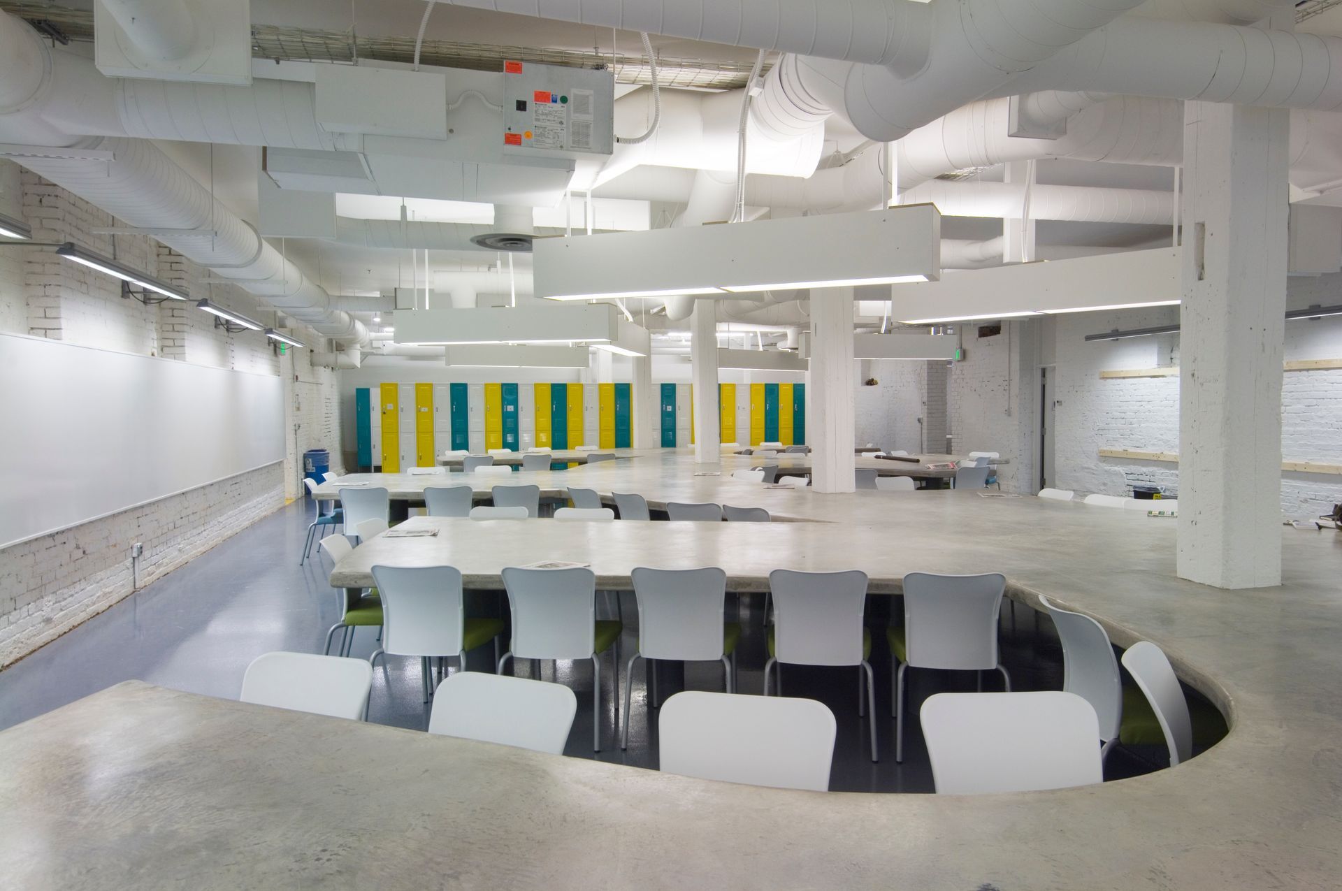 Open, modern room with curved tables, white chairs, and ceiling fixtures. Yellow and green lockers in the background.