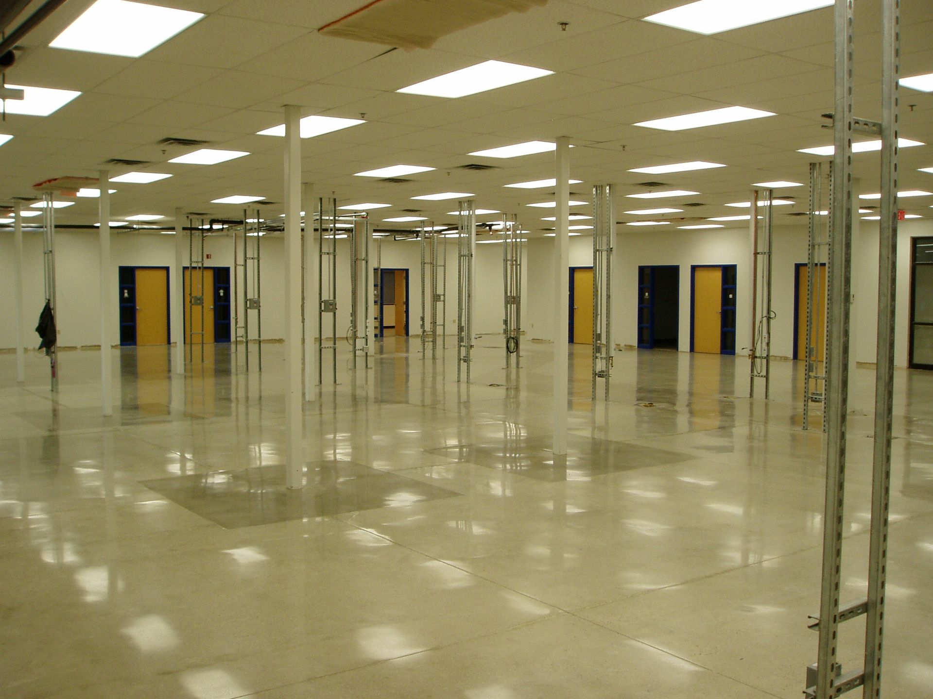 Empty, brightly lit commercial space with support beams, reflective flooring, and several closed doors in the back.