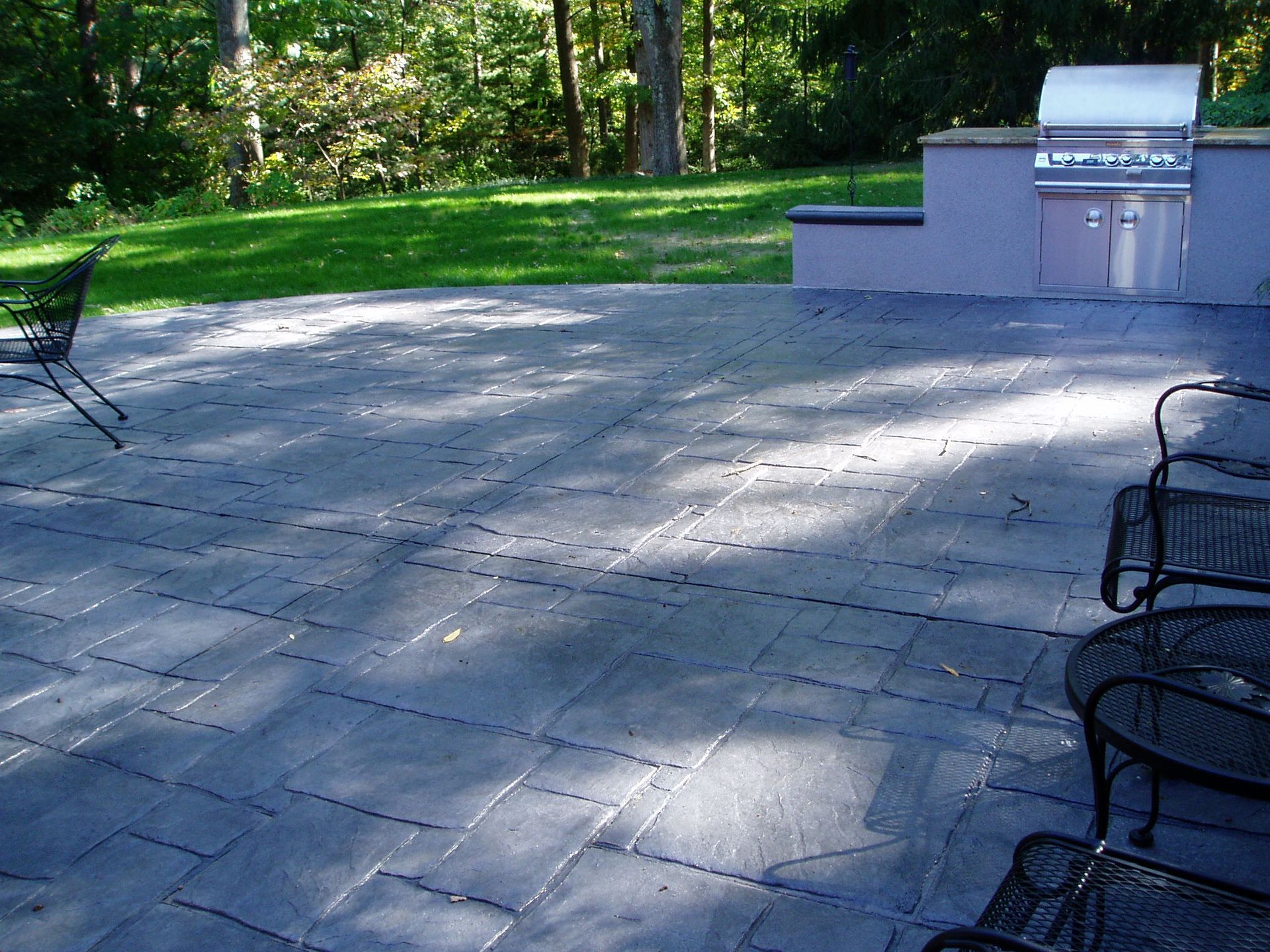 Patio with dark gray pavers, grill, and metal chairs, in a grassy backyard.