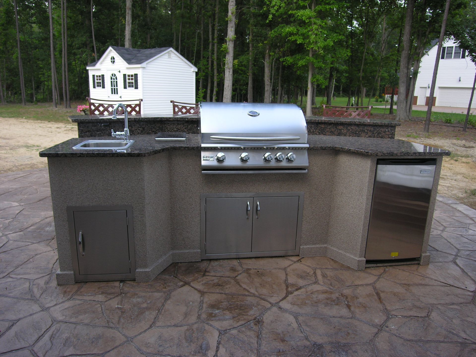 Outdoor kitchen with stainless steel grill, sink, and refrigerator on a stone patio.