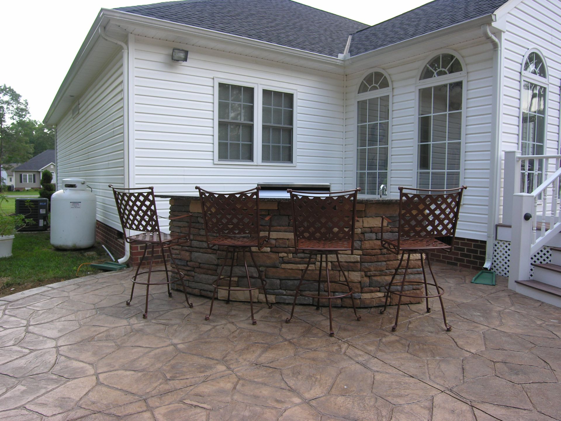 Outdoor bar with stone facade, metal chairs, built-in grill, and stamped concrete patio next to a house.
