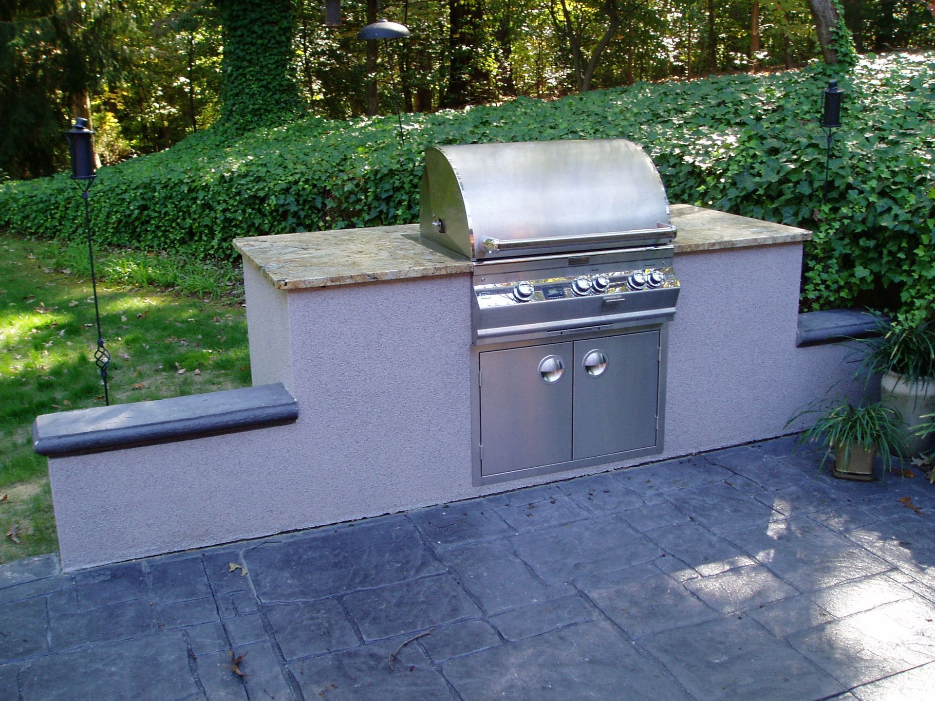 Outdoor built-in grill with stainless steel features, set on a stone patio with greenery backdrop.