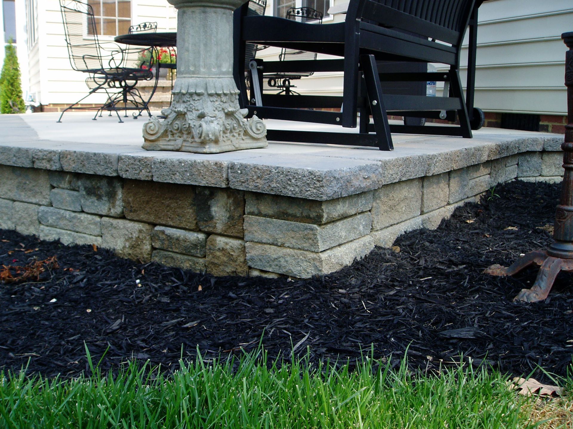 Stone retaining wall supporting a patio with a bench, garden, and grass.