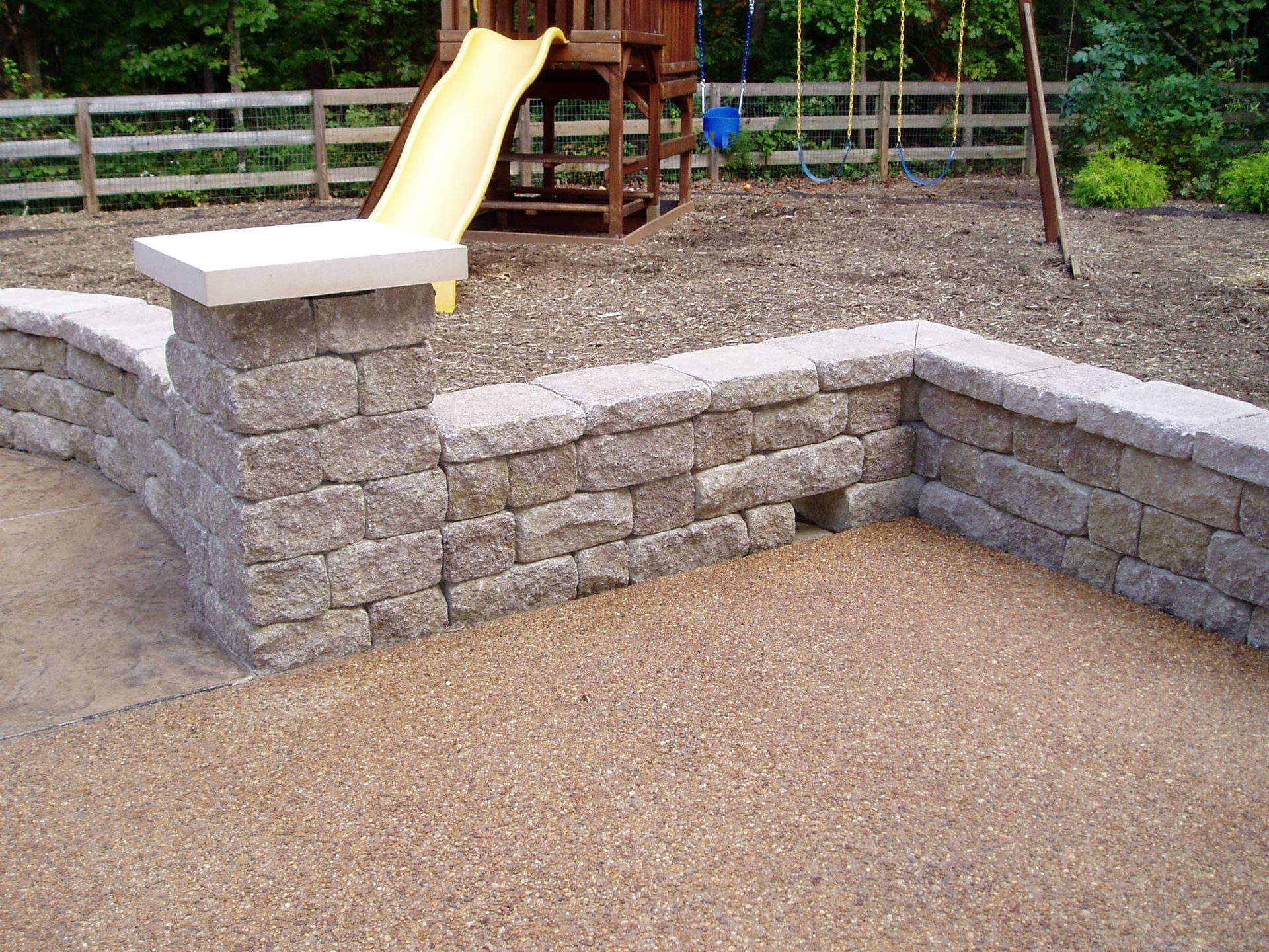 Low retaining wall made of gray blocks with a tan play area and a yellow slide in the background.