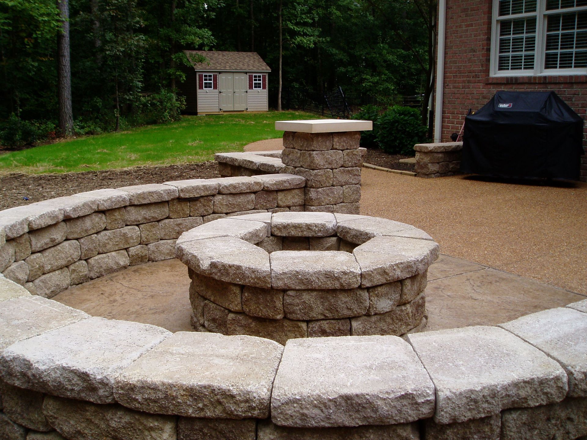 Stone fire pit with surrounding walls on a gravel patio, near a brick house and a shed.