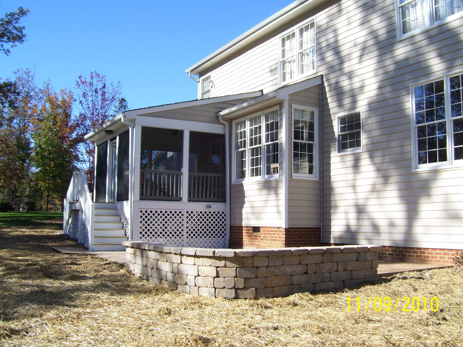 A screened-in porch with a stone retaining wall and house in the background on a sunny day.