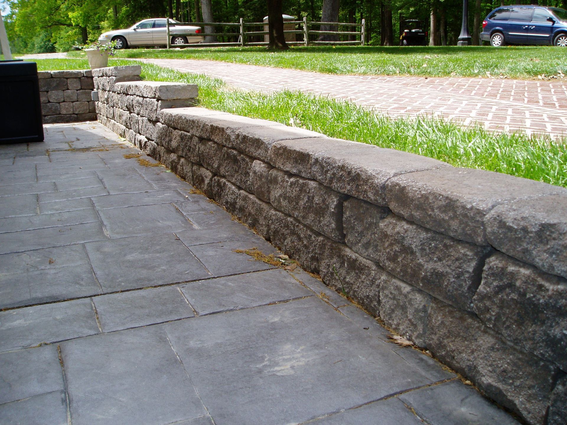 Stone wall along a patio with a grassy area in the background. A car is visible in the distance.
