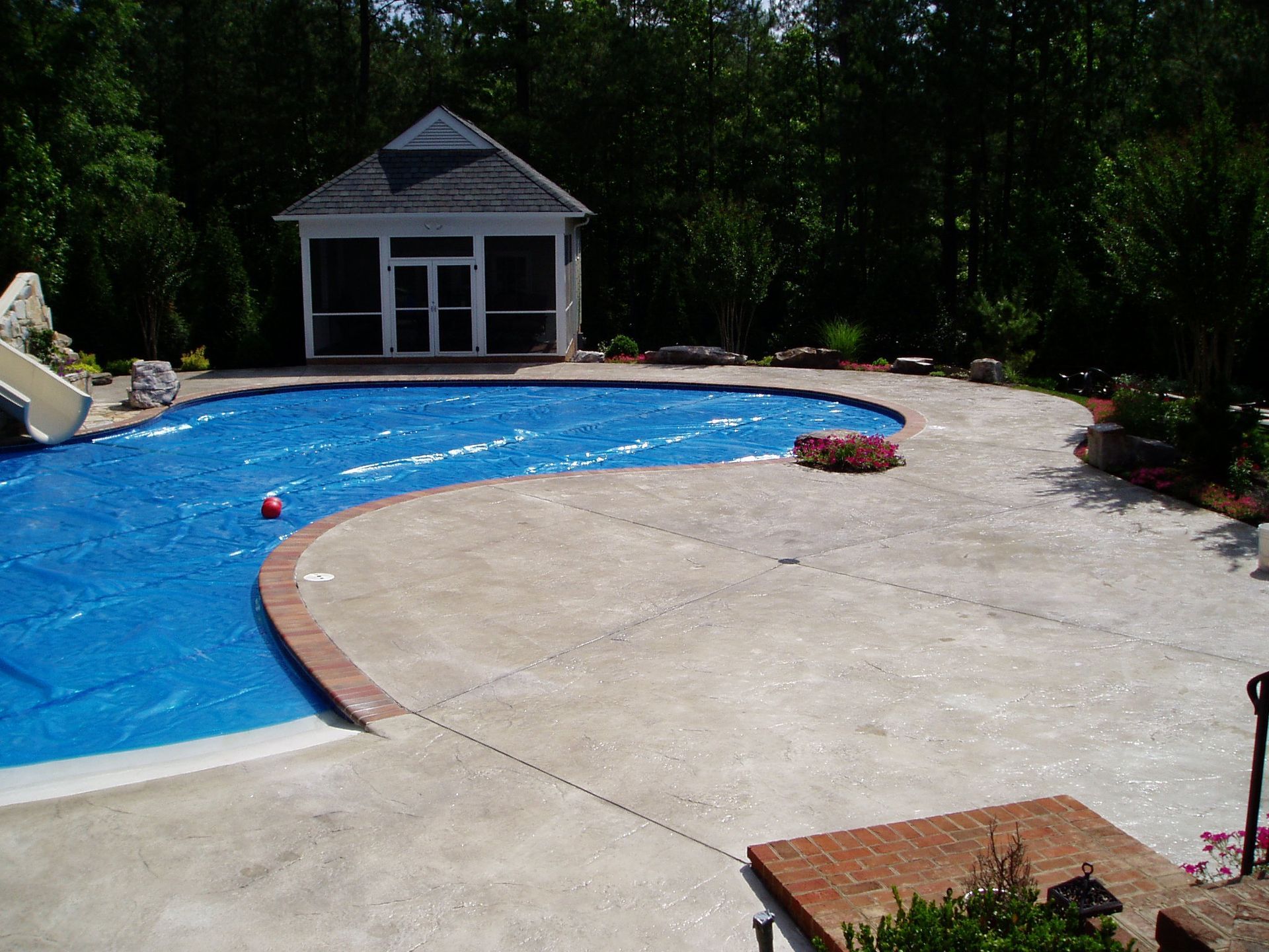 Blue-covered swimming pool with a gazebo, surrounded by a concrete patio and greenery.