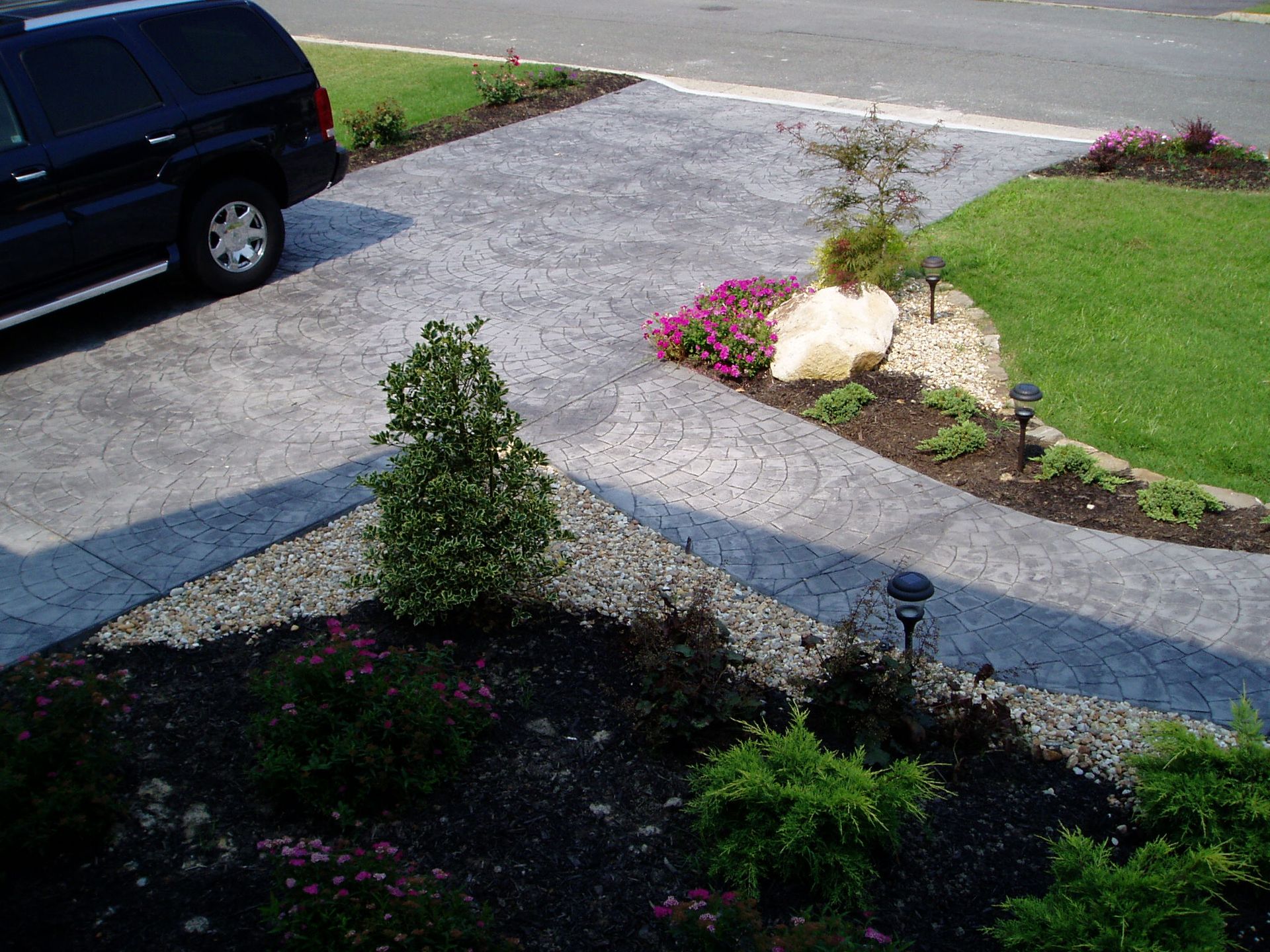 Black SUV parked on gray patterned concrete driveway. Landscaped yard with plants and mulch.