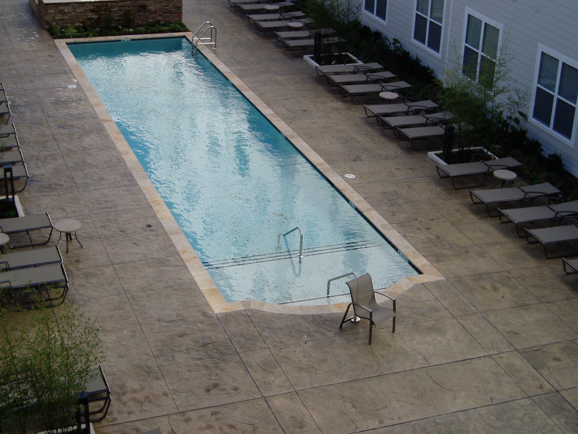 A rectangular swimming pool surrounded by a concrete patio with lounge chairs.