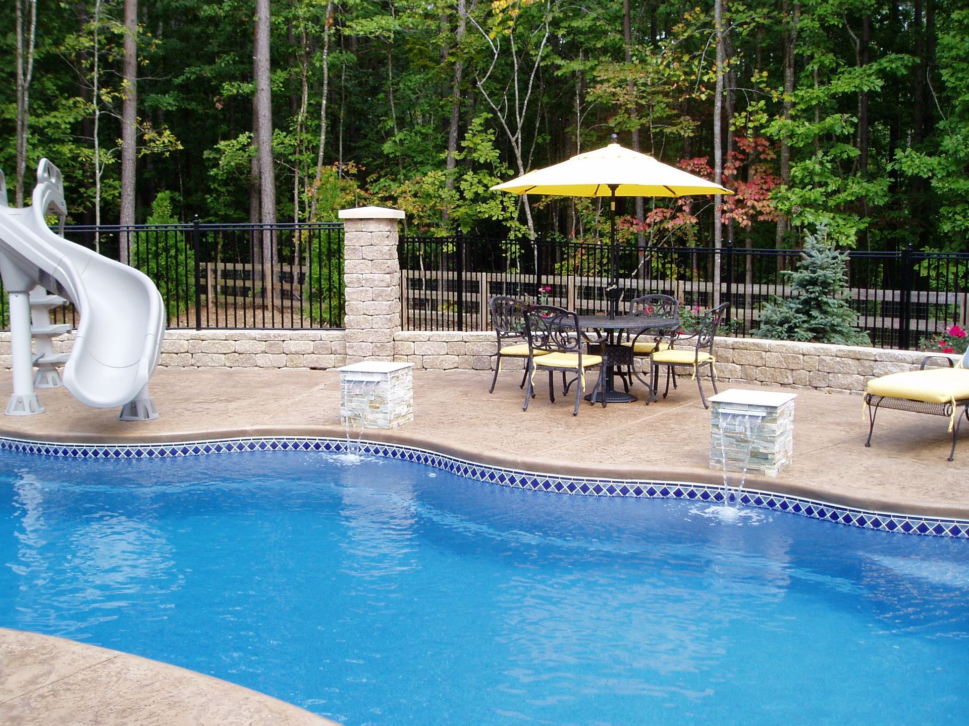 Swimming pool with slide, patio furniture under yellow umbrella, stone wall and forest background.