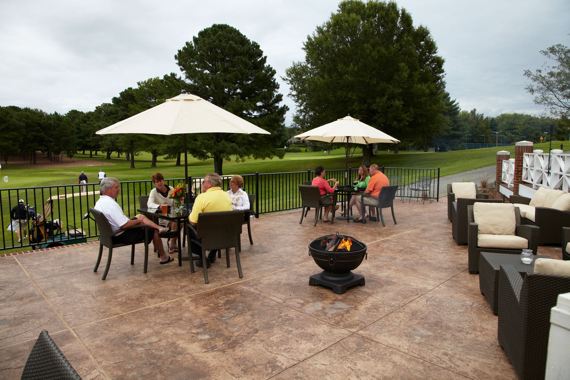 Patio with people at tables, overlooking a golf course. Fire pit, umbrellas, trees.