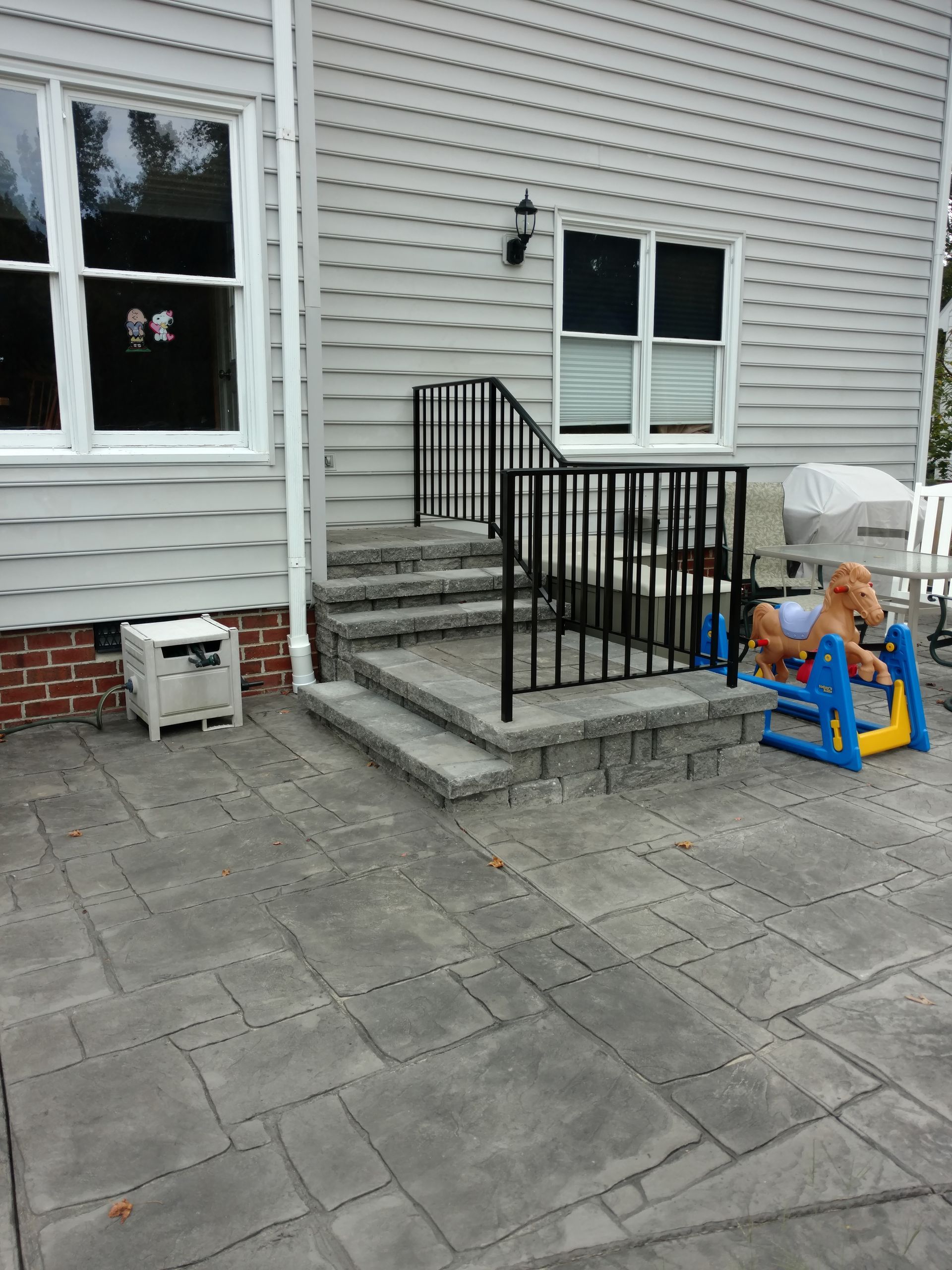Grey stone patio with steps leading to a raised area with black railing. Siding on the house is grey.