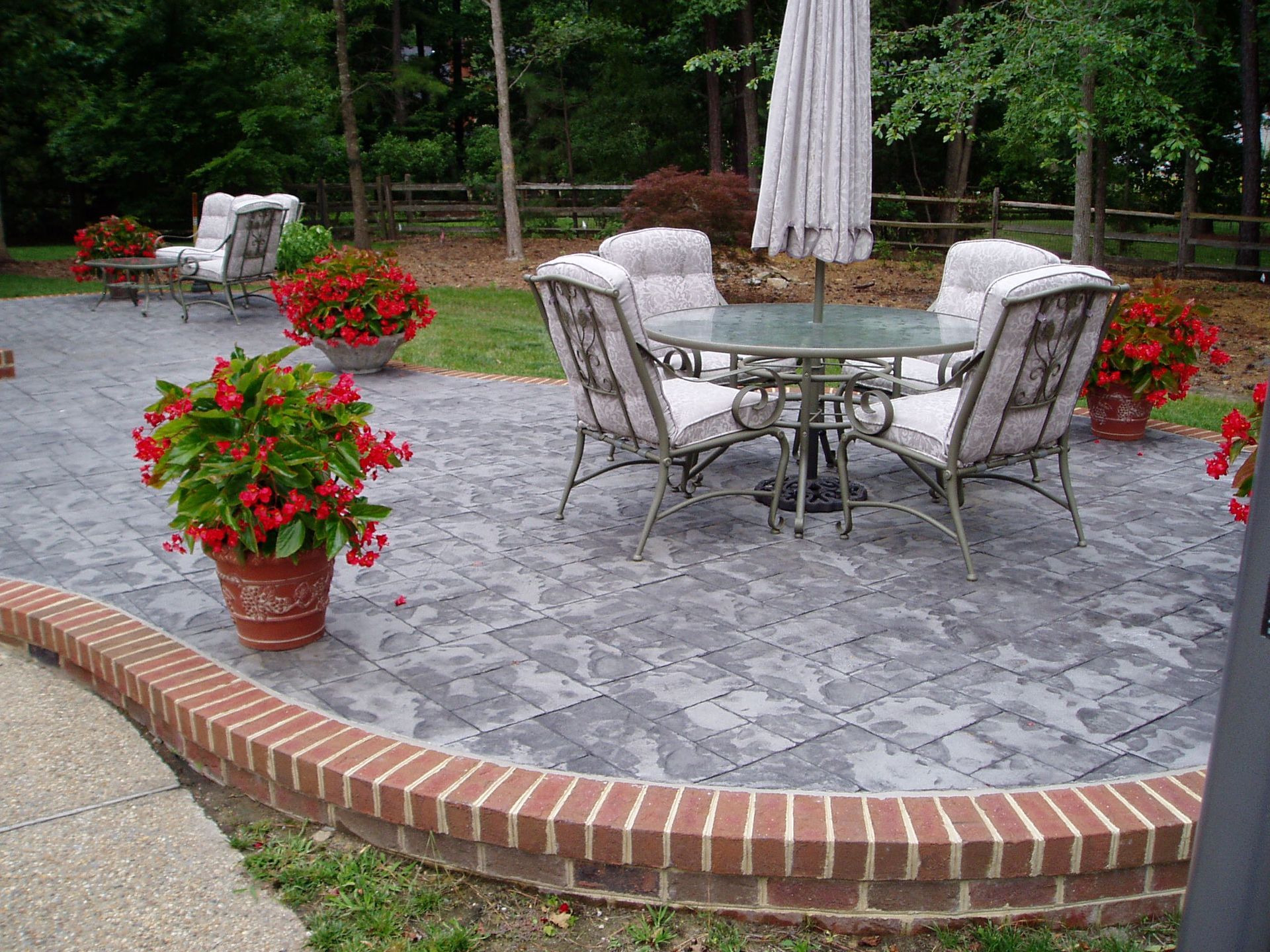Patio with round table, chairs, potted red flowers, and brick edging.