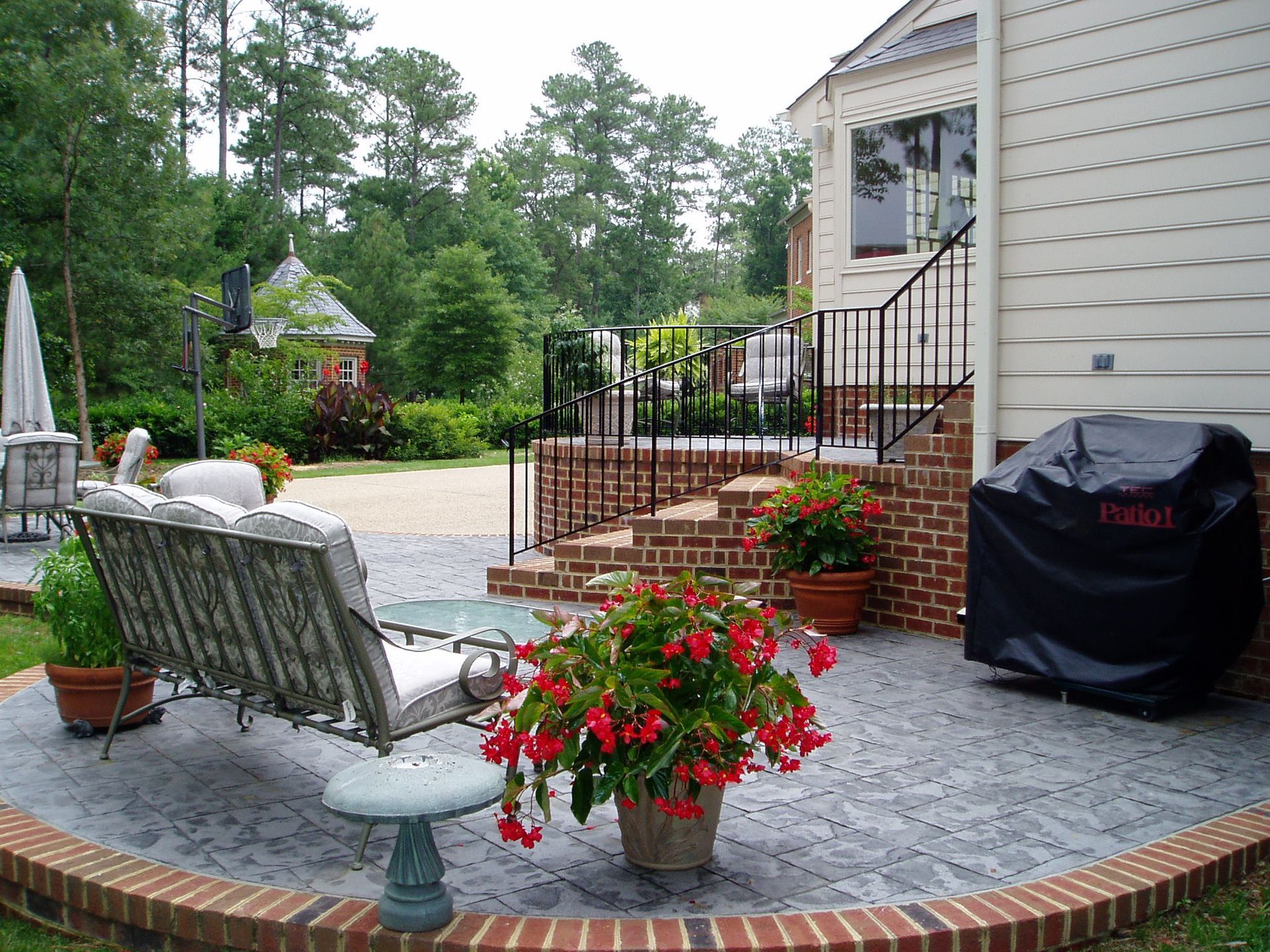 Patio with brick border, seating, and potted red flowers next to a house with steps.