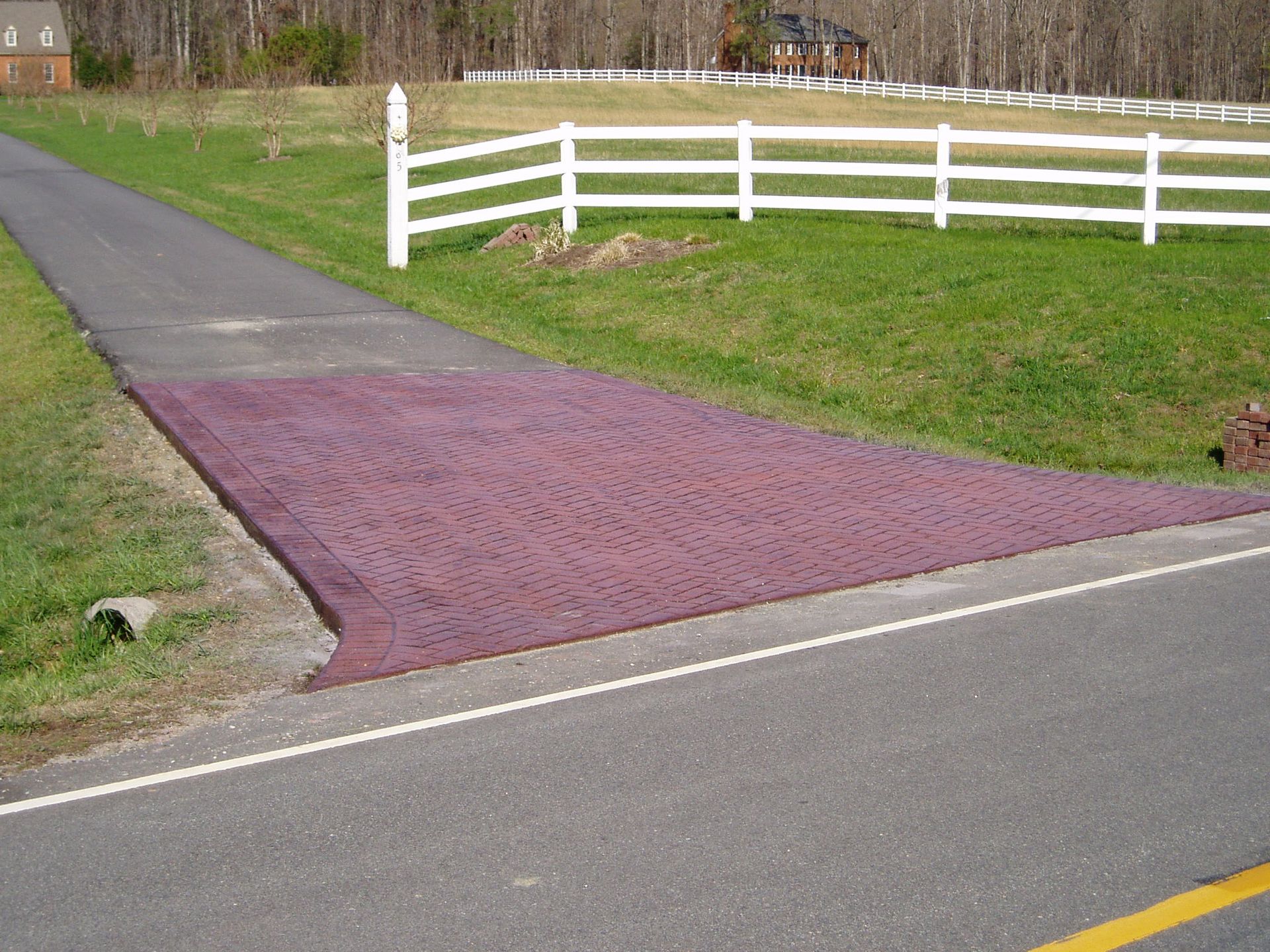 Brick driveway crossing road to a rural property with white fence, green grass, and trees.