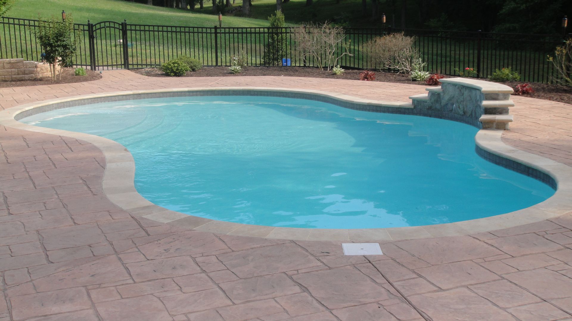 Pool with blue water and stone deck. Waterfall steps and black fence in background.
