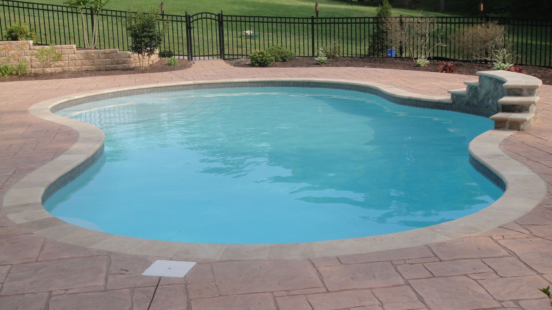 Swimming pool with blue water and stone patio. Steps lead up to a garden area behind a black fence.